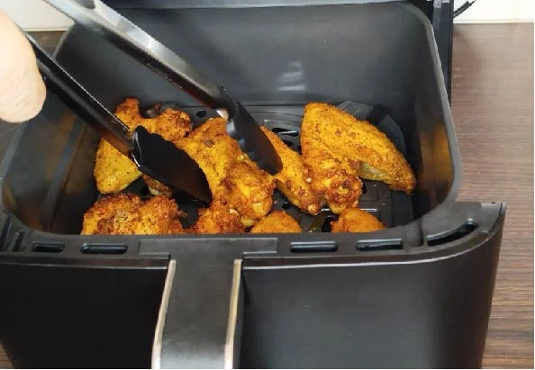 Des ailes de poulet croustillantes dans une friteuse à air, manipulées avec des pinces pour une cuisson parfaite.