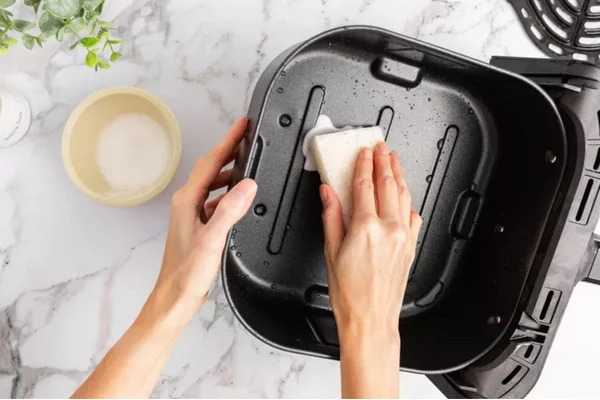 Une main nettoie le panier d'une friteuse à air avec une éponge, illustrant l'importance de l'entretien.
